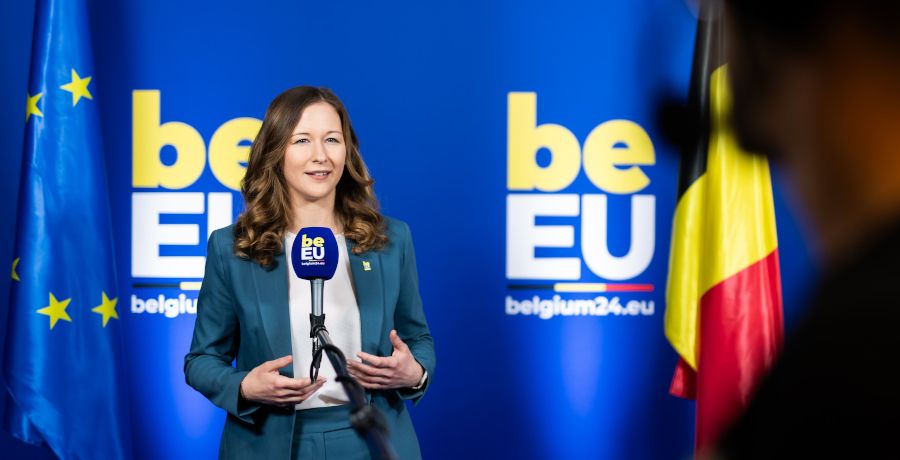 Staatssekretärin Claudia Plakolm steht vor einem Mikrofon, im Hintergrund EU- und Belgien-Fahne