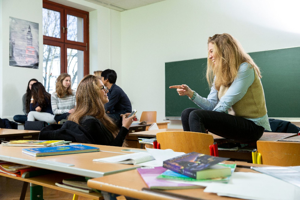 Zwei Schülerinnen sprechen miteinander in einem Klassenraum, im Hintergrund sind weitere Personen zu sehen