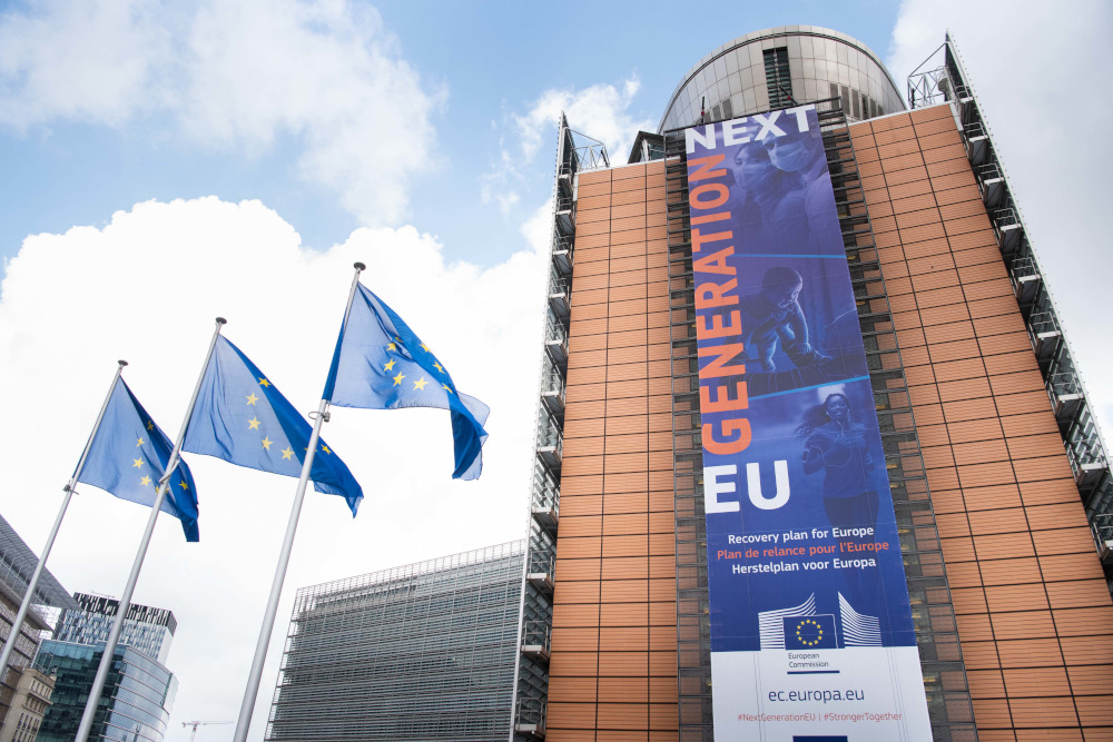 Das Banner "Recovery Plan für Europa" hängt auf der Front des Berlaymont Gebäude - im Vordergrund sind drei wehende EU-Flaggen zu sehen