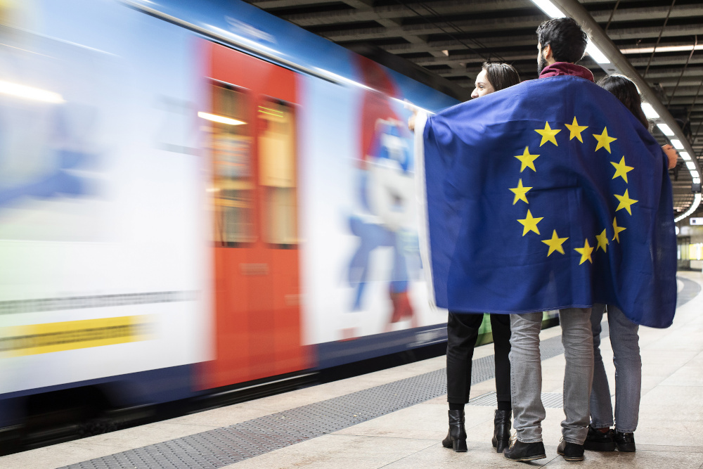 Drei Personen stehen im Kreis an einem Bahnsteig und sind von einer EU-Flagge umhüllt. Daneben ist ein vorbei fahrender Zug zu sehen.